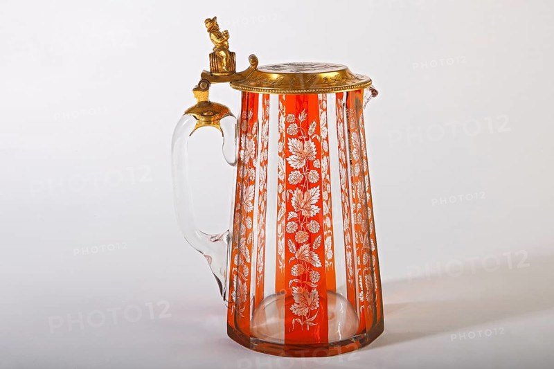 Decorative old beer stein made of crystal glass with hop ornaments and brass lid with beer drinking figure sitting on beer barrel