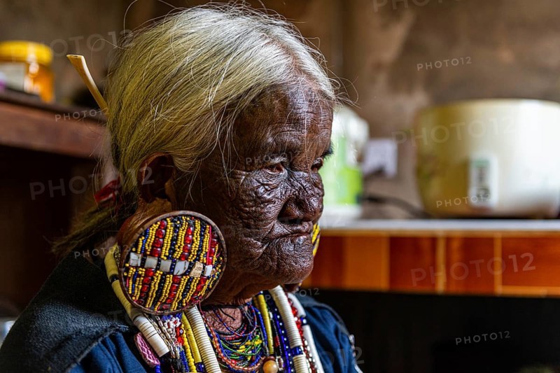 Chin Kaang hill-tribe woman with traditional facial tattoo and elaborate tribal earrings playing a flute with her nose