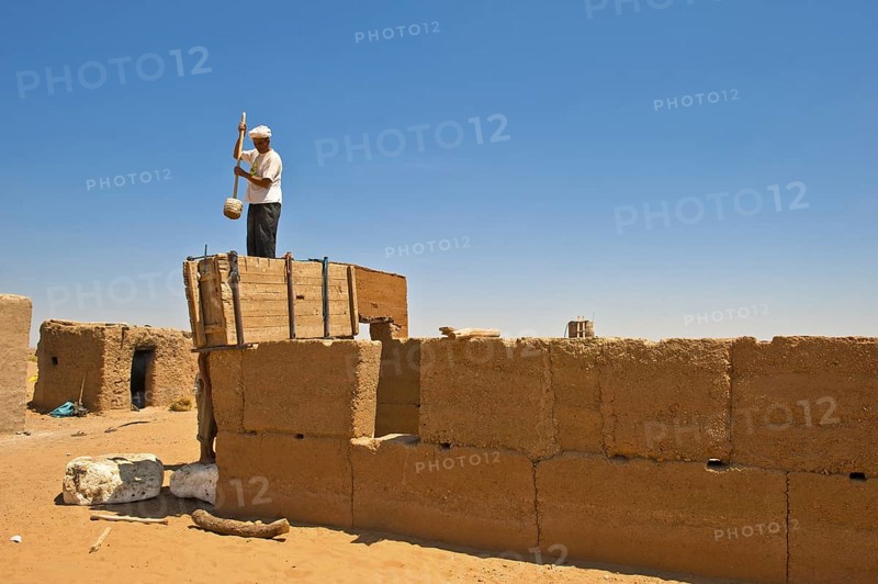 Elderly man wearing a white turban using a wooden tool to compact clay into a wooden casing for the construction of a wall made of rammed earth