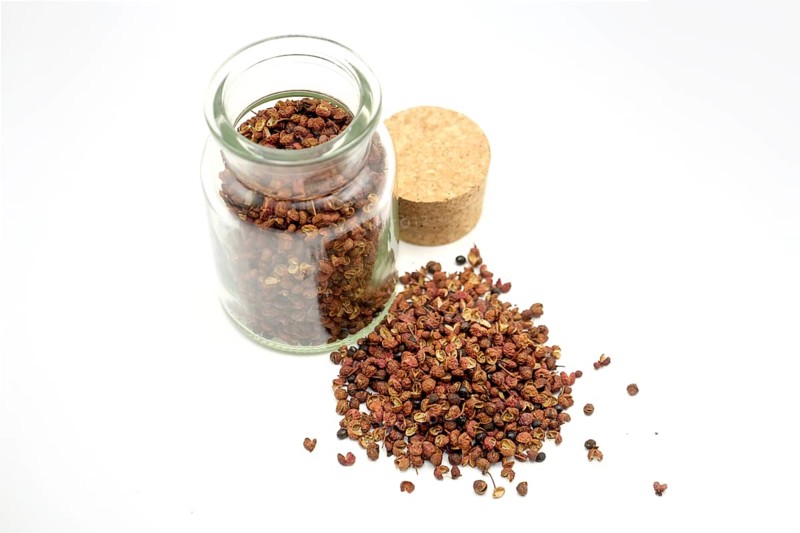 Sichuan peppercorns beside and in a 150ml glass spice jar against a white background
