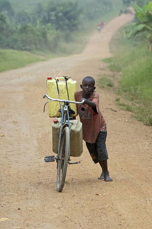 Boy pushing bicycle laden with full water containers along dusty road