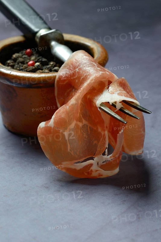 A slice of coppa on fork and clay pot with peppercorns