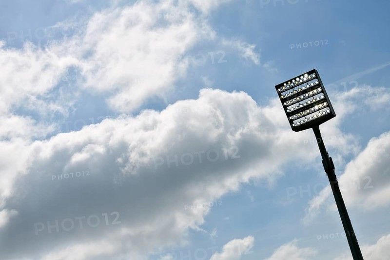 Floodlight mast switched on in front of blue sky with clouds