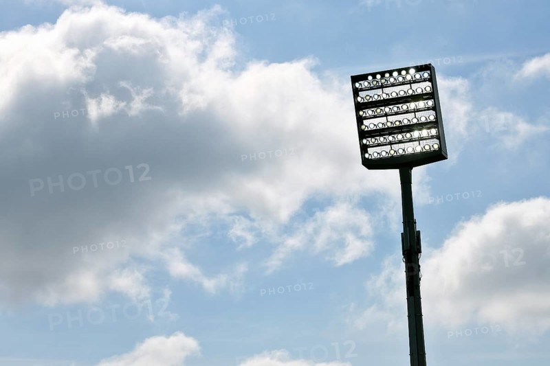 Floodlight mast switched on in front of blue sky with clouds