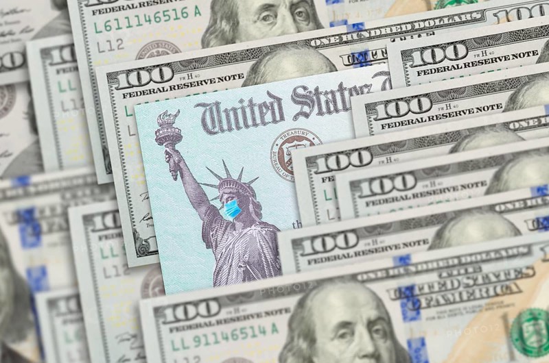 United states IRS stimulus check with statue of liberty wearing medical face mask resting on hundred dollar bills