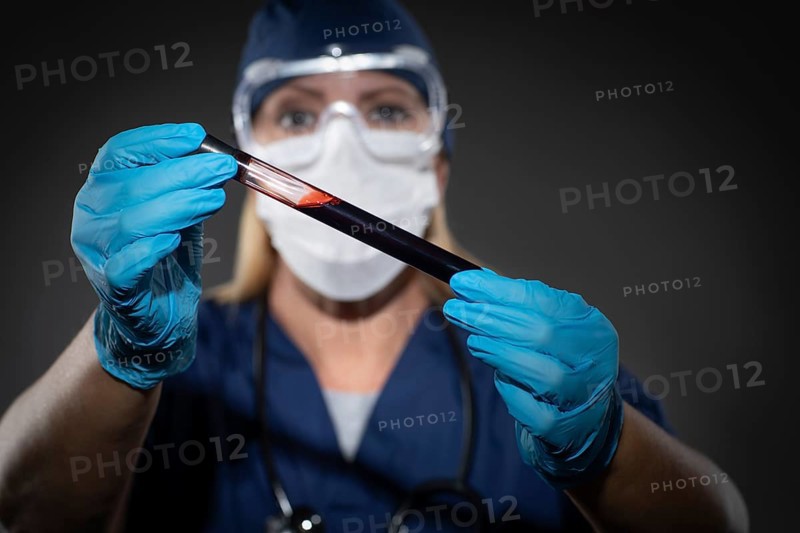 Female Lab Worker Wearing Medical Face Mask Holds Test Tube of Blood Against Dark Background