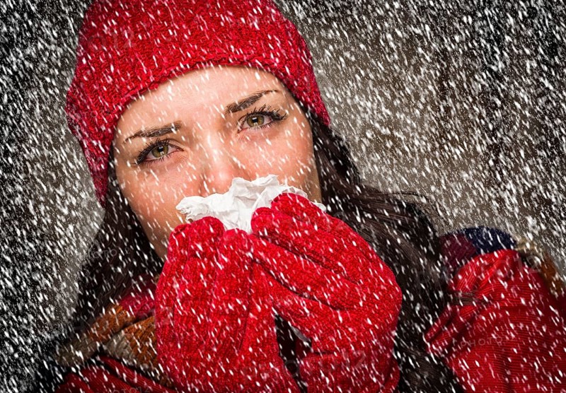Sick mixed-race woman wearing winter hat and gloves blowing her sore nose with a tissue in the snow