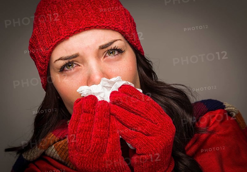 Sick mixed-race woman wearing winter hat and gloves blowing her sore nose with a tissue