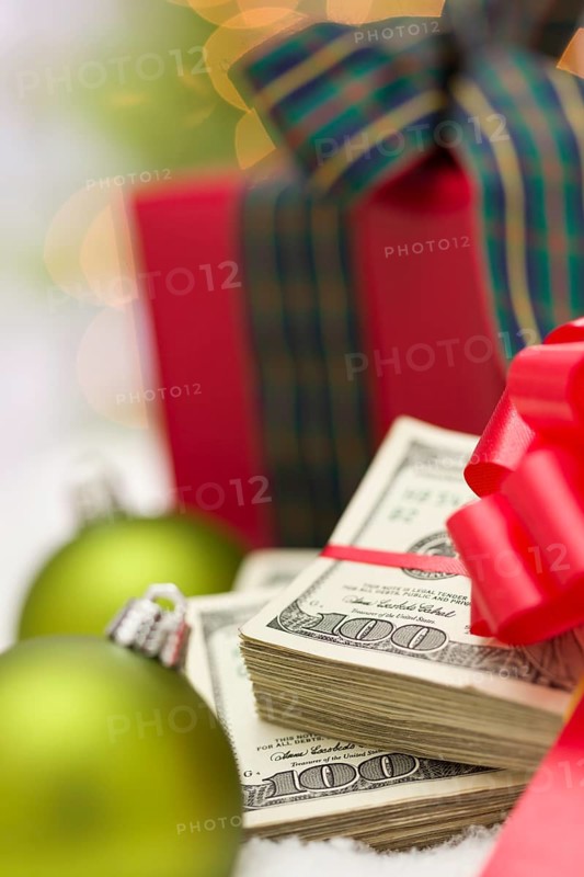 Stack of one hundred dollar bills with red bow near green christmas ornaments and wrapped gift box on snow flakes