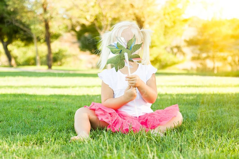 Cute little girl sitting in grass blowing on pinwheel toy