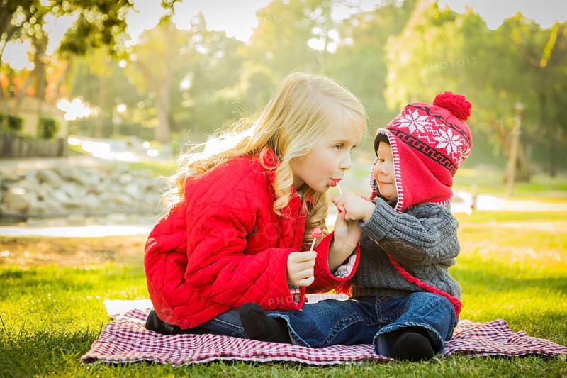 Little girl with her baby brother wearing winter coats and hats sharing a lollipop outdoors at the park