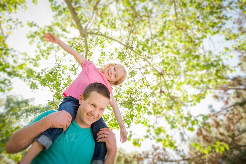 Cute young girl rides piggyback on her dads shoulders outside at the park