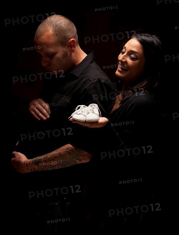 Fun mixed-race couple holding new white baby shoes against a black background under dramatic lighting