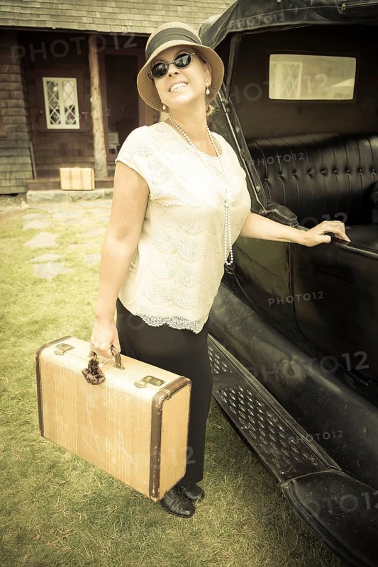 Happy 1920s dressed girl holding suitcase next to vintage car and cabin
