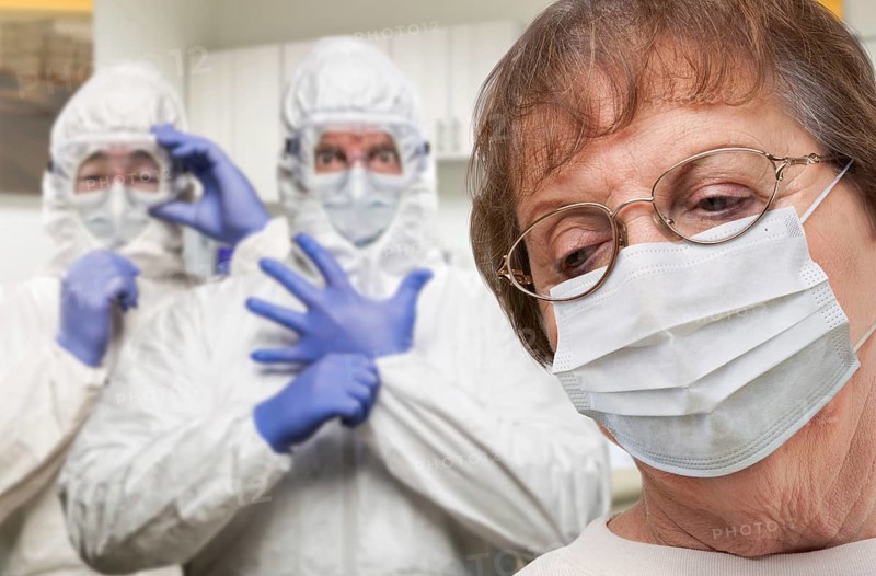 Senior adult woman wearing medical face mask in doctor office with nurses in HAZMAT suits behind