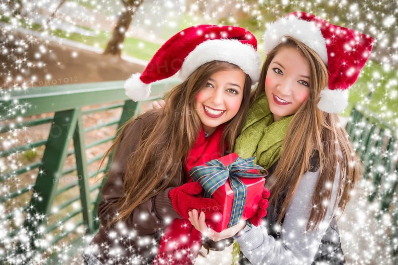 Two attractive festive smiling mixed-race women wearing christmas santa hats holding a bow wrapped gift outside with snow flakes border
