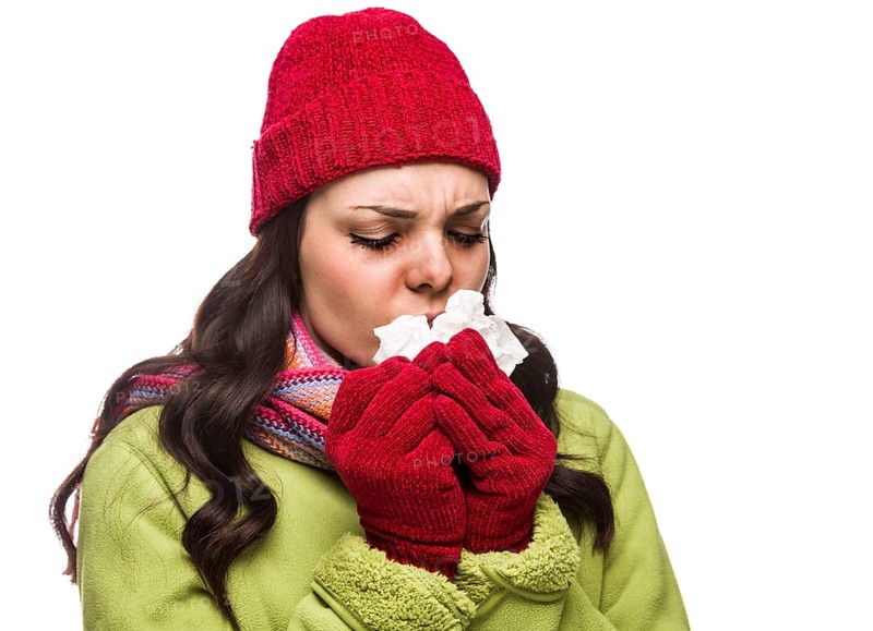 Sick mixed-race woman wearing winter hat and gloves blowing her sore nose with a tissue isolated on white background
