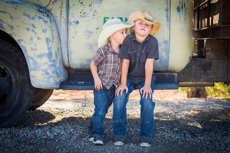 Two young boys wearing cowboy hats leaning against an antique truck in a rustic country setting