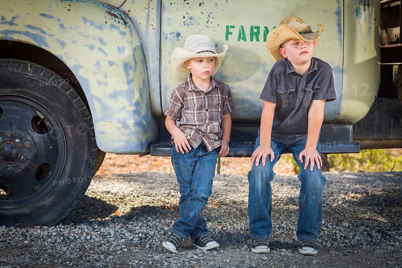 Two young boys wearing cowboy hats leaning against an antique truck in a rustic country setting