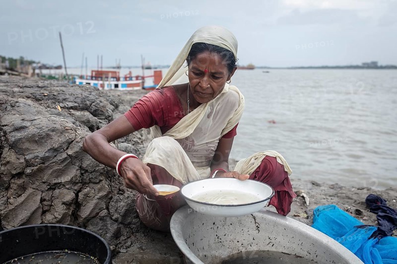 A woman sieves river water from wash bowls on a muddy embankment