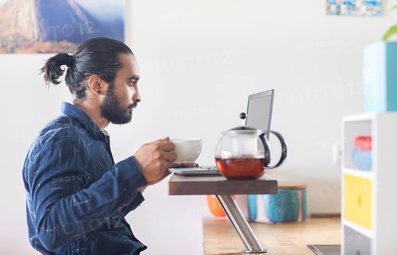 Young man with ponytail and checkered shirt drinking tea at home and working