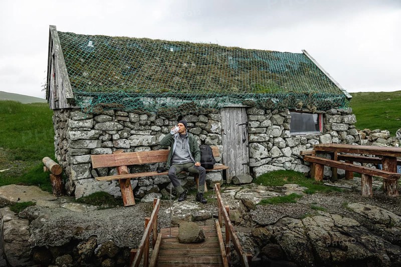 Hiker drinking water on a bench in front of a stone hut
