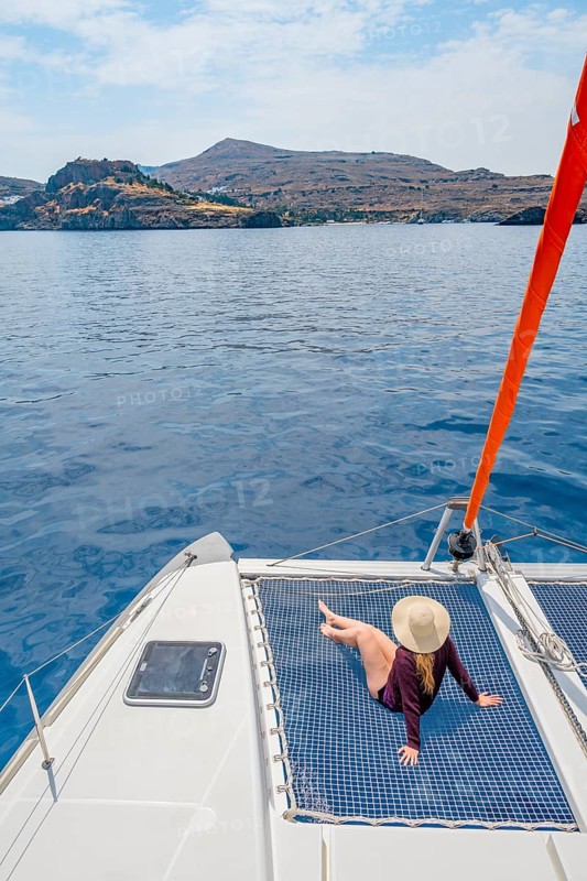 Young woman with hat sitting in the net of a sailing catamaran