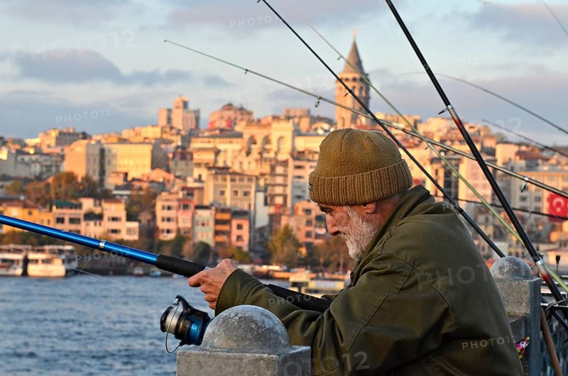 Old man with cap fishing at the Golden Horn on Galata Bridge with view the Galata Tower at sunset