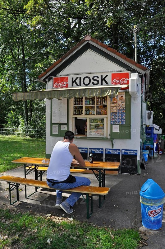 Man in white vest and jeans sits on bench in front of kiosk