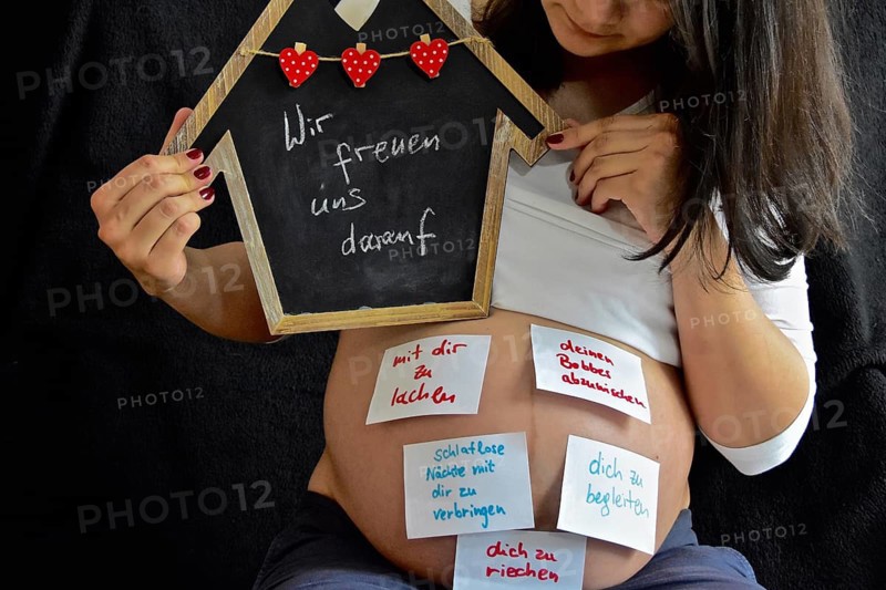 Expectant with wish slips pasted on her stomach holds up black board with saying Wir freuen uns drauf