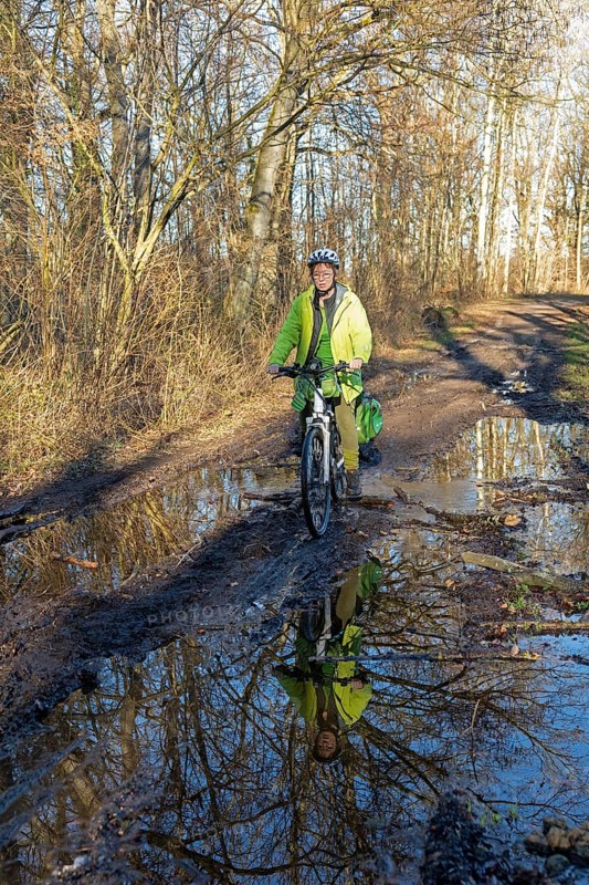 Woman cycling with e-bike over muddy forest path and through puddles