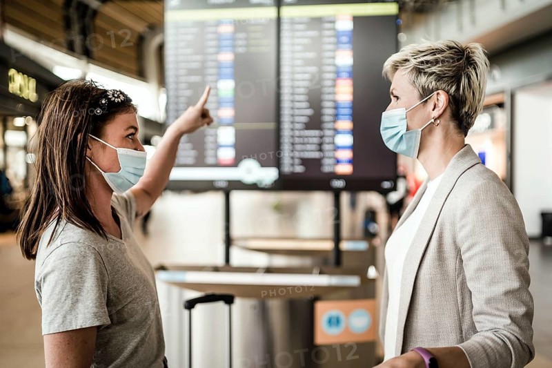 Two travelling women wearing protective masks discussing by flight information board at the Faro airport