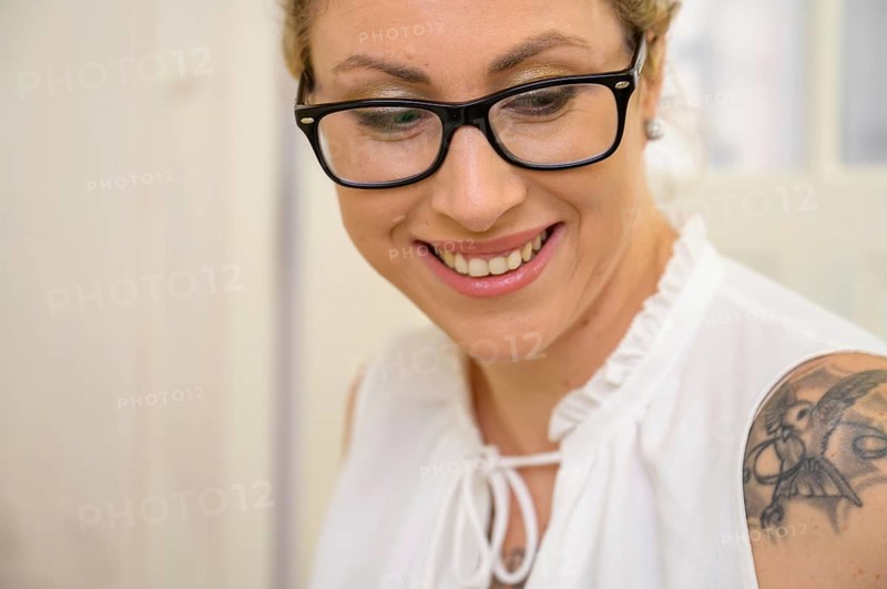Portrait of a pretty young woman with blonde hair tied back and large black glasses looks down with a smile and beautiful teeth. The sleeveless white blouse reveals a tattooed shoulder