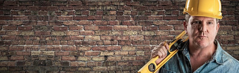 Male contractor in hard hat holding level in front of old brick wall banner with copy space