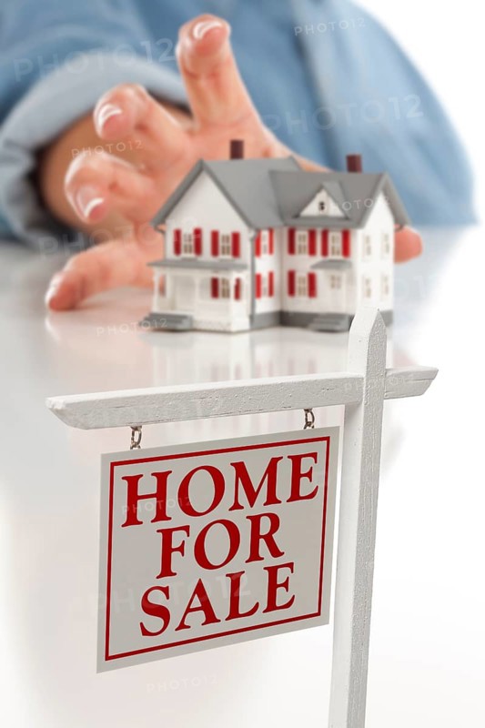 Real estate sign in front of womans hand reaching for model house on a white surface