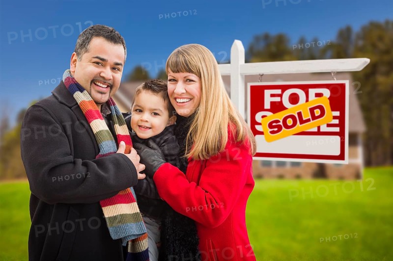 Warmly dressed young mixed-race family in front of sold home for sale real estate sign and house