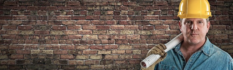 Male contractor in hard hat holding construction plans in front of old brick wall banner with copy space