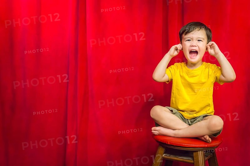 mixed-race boy with his fingers in his ears sitting on stool in front of red curtain