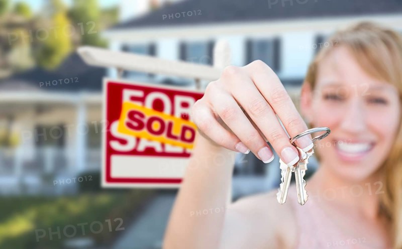 Excited woman holding house keys and sold for sale real estate sign in front of nice new home