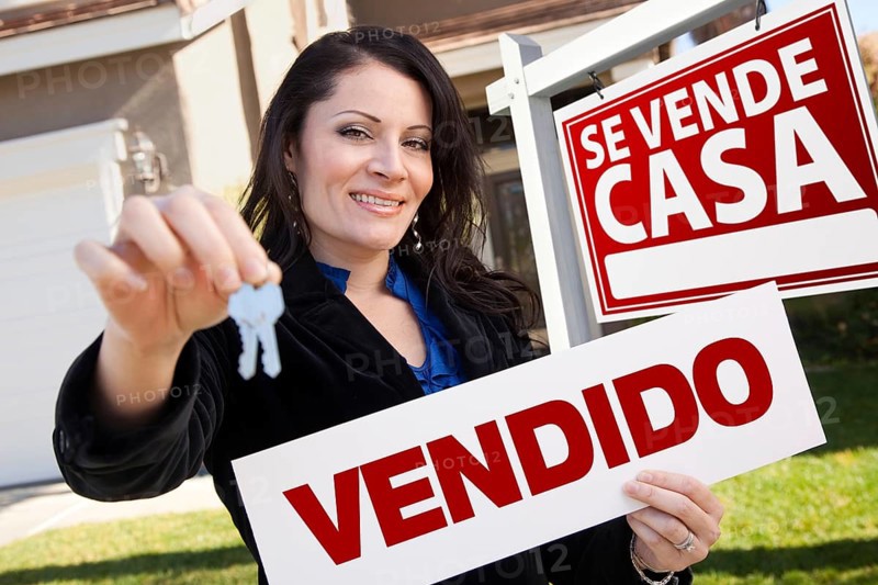 Happy attractive hispanic woman holding vendido real estate sign and keys in front se vende casa real estate sign and house