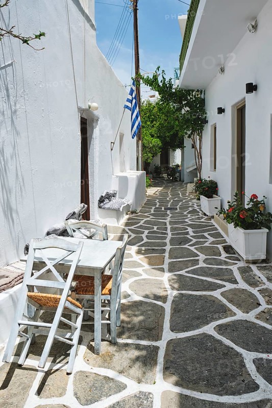 Picturesque narrow street with traditional whitewashed houses with greek flag and cafe tables in Naousa town in famous tourist attraction Paros island