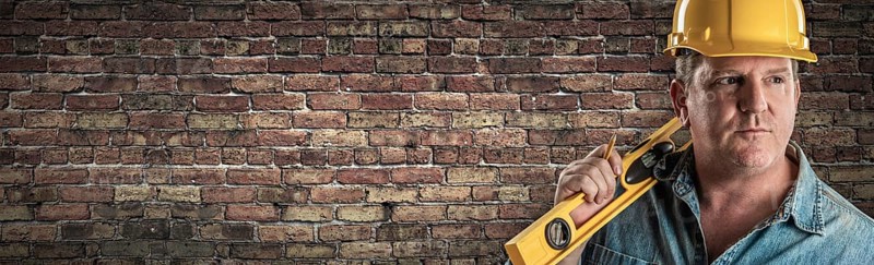 Male contractor in hard hat holding level in front of old brick wall banner with copy space