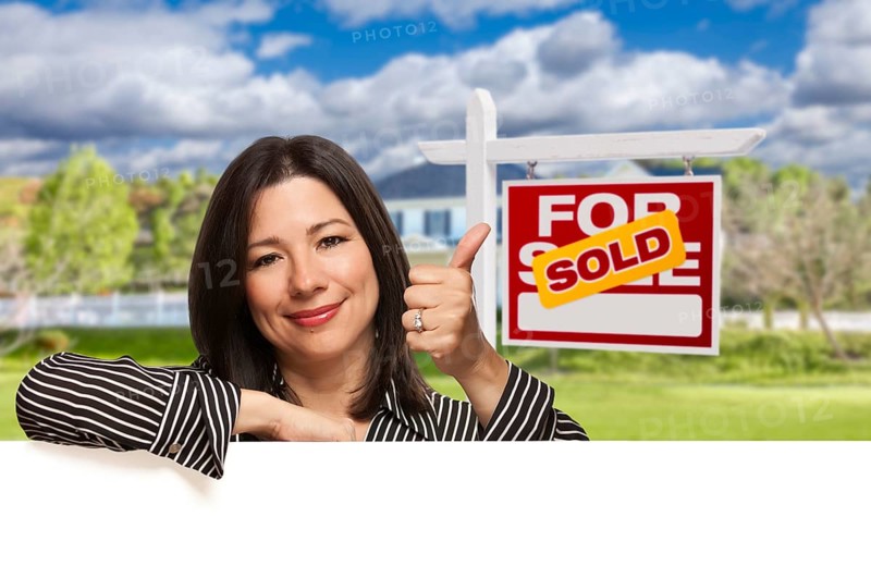Pretty hispanic woman leaning on white with thumbs up in front of beautiful house and sold for sale real estate sign