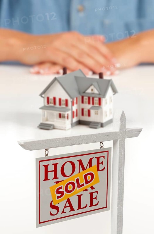 Womans folded hands behind model house and sold home for sale real estate sign in front on white surface