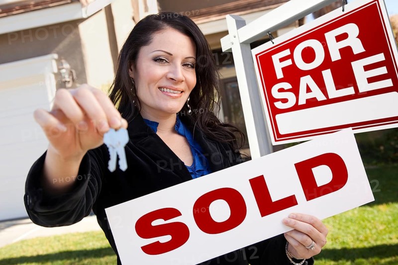 Happy attractive hispanic woman holding sold real estate sign and keys in front for sale real estate sign and house