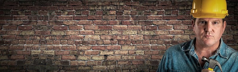 Male contractor in hard hat holding hammer in front of old brick wall banner with copy space