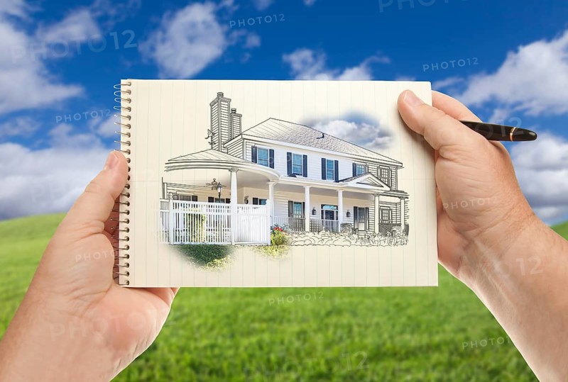 Male hands holding pen and pad of paper with house drawing over empty grass field and sky