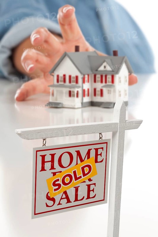 Sold real estate sign in front of womans hand reaching for model house on a white surface