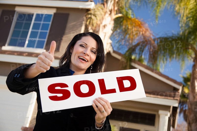 Happy attractive hispanic woman with thumbs up holding sold sign in front of house