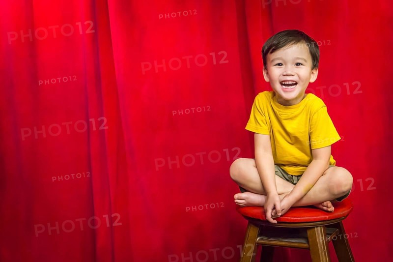 Laughing mixed-race boy sitting on stool in front of red curtain